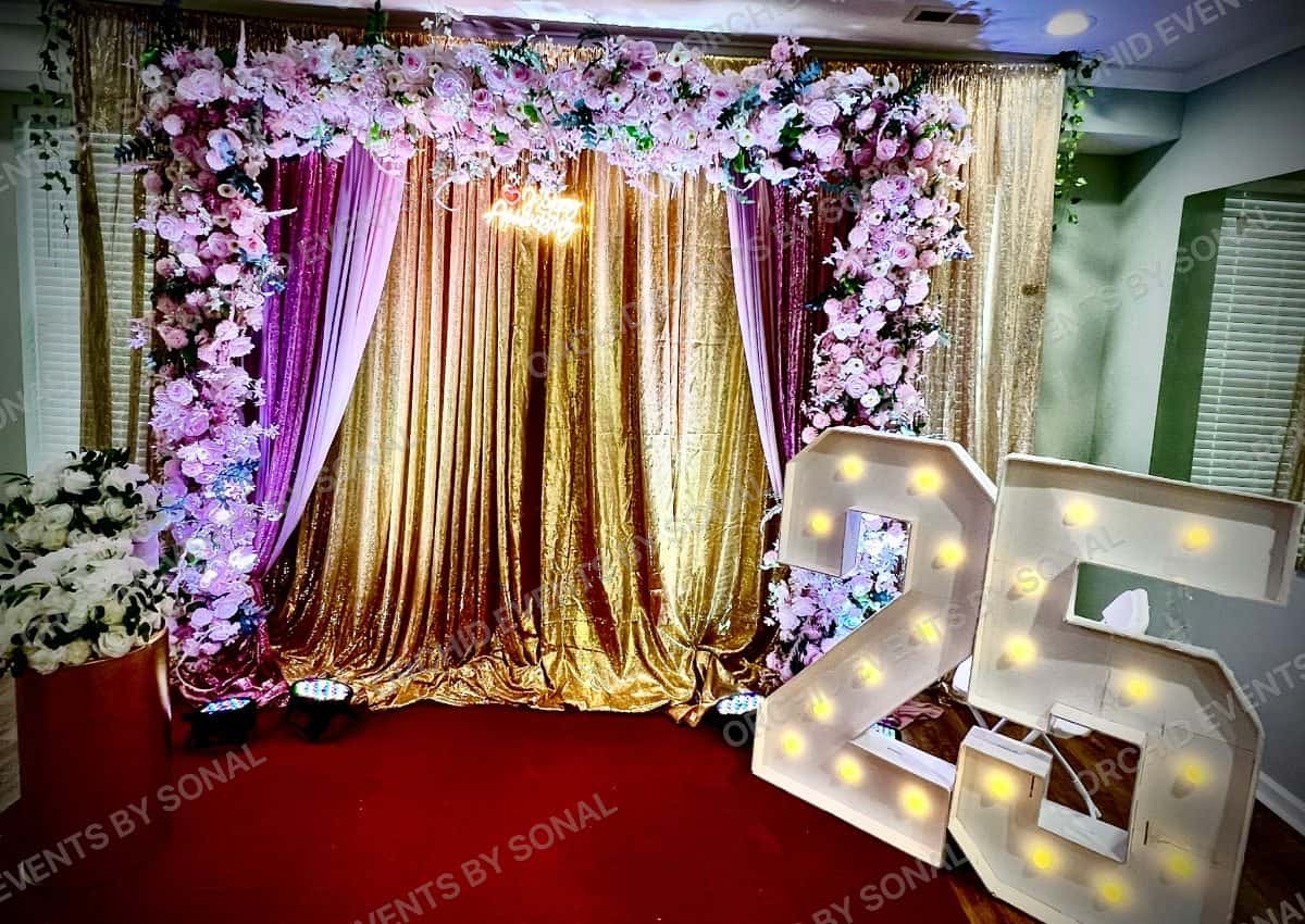Gold sequin anniversary backdrop with a lit 25 marquee and a blossom arch