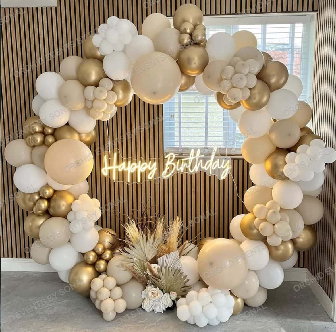 Cream and gold balloon arch around a neon Happy Birthday sign, with pampas grass at the base