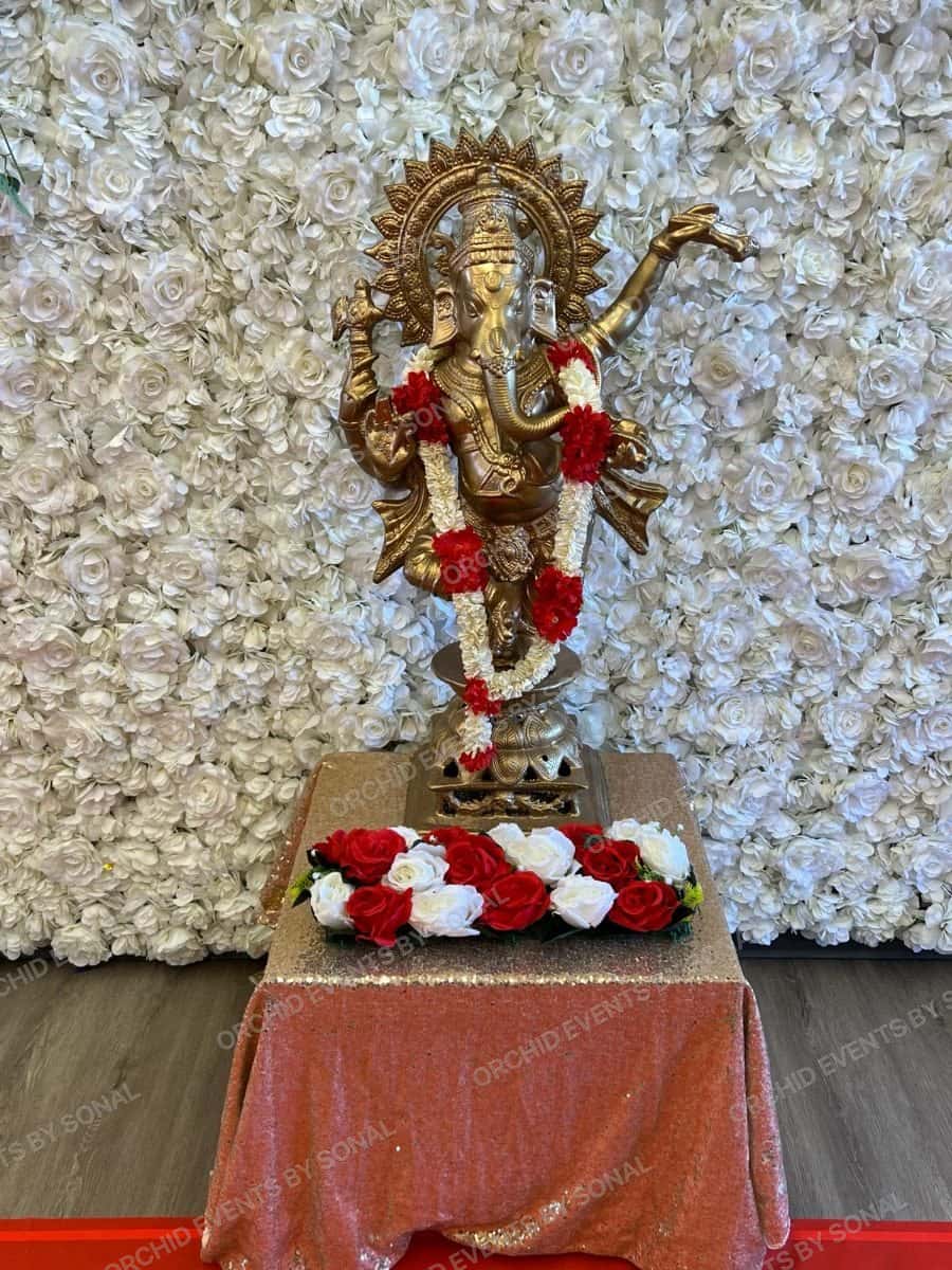 Brass Ganesh on a white rose flower wall with a red and white garland