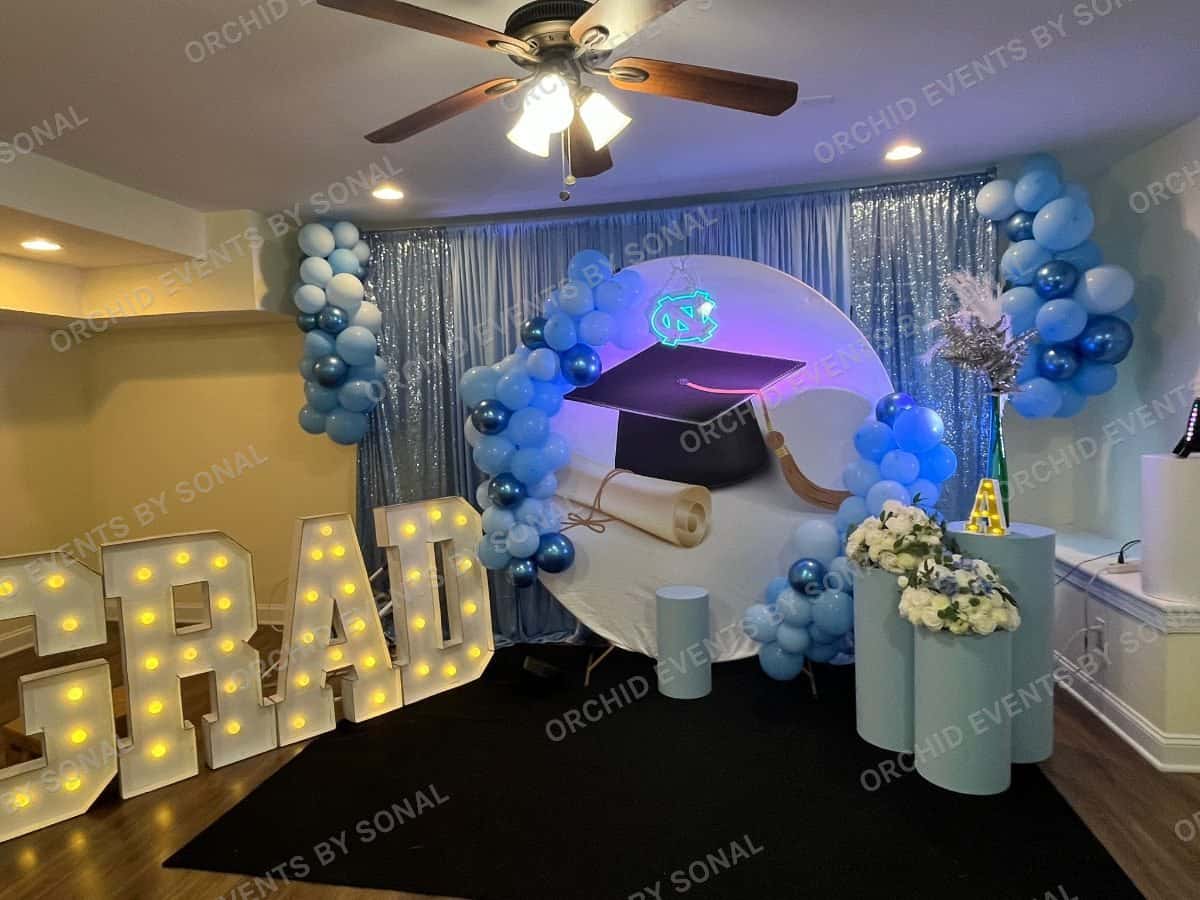 Blue balloon arch and mortarboard over lit GRAD letters in a pale function room