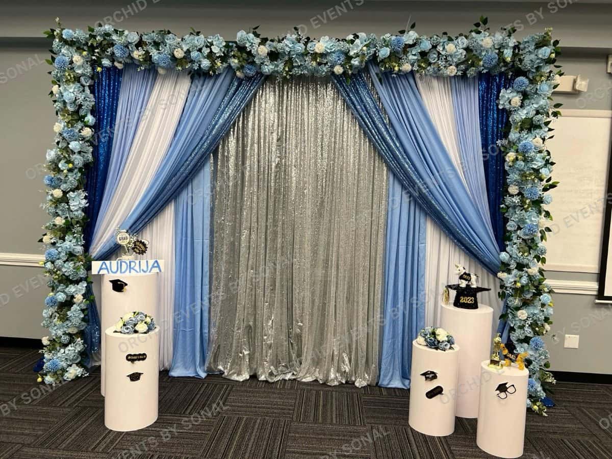 Graduation setup in pale blue and silver drape with a white blossom arch and plinths