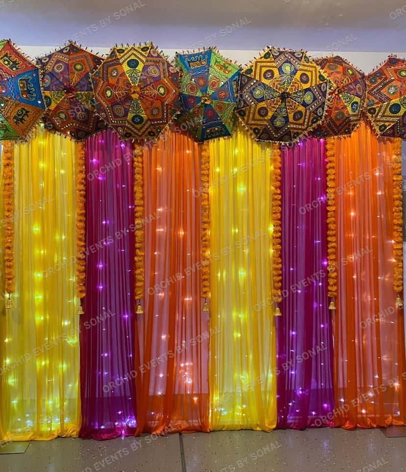 Row of embroidered Rajasthani umbrellas above fairy-lit curtains in yellow, magenta and orange